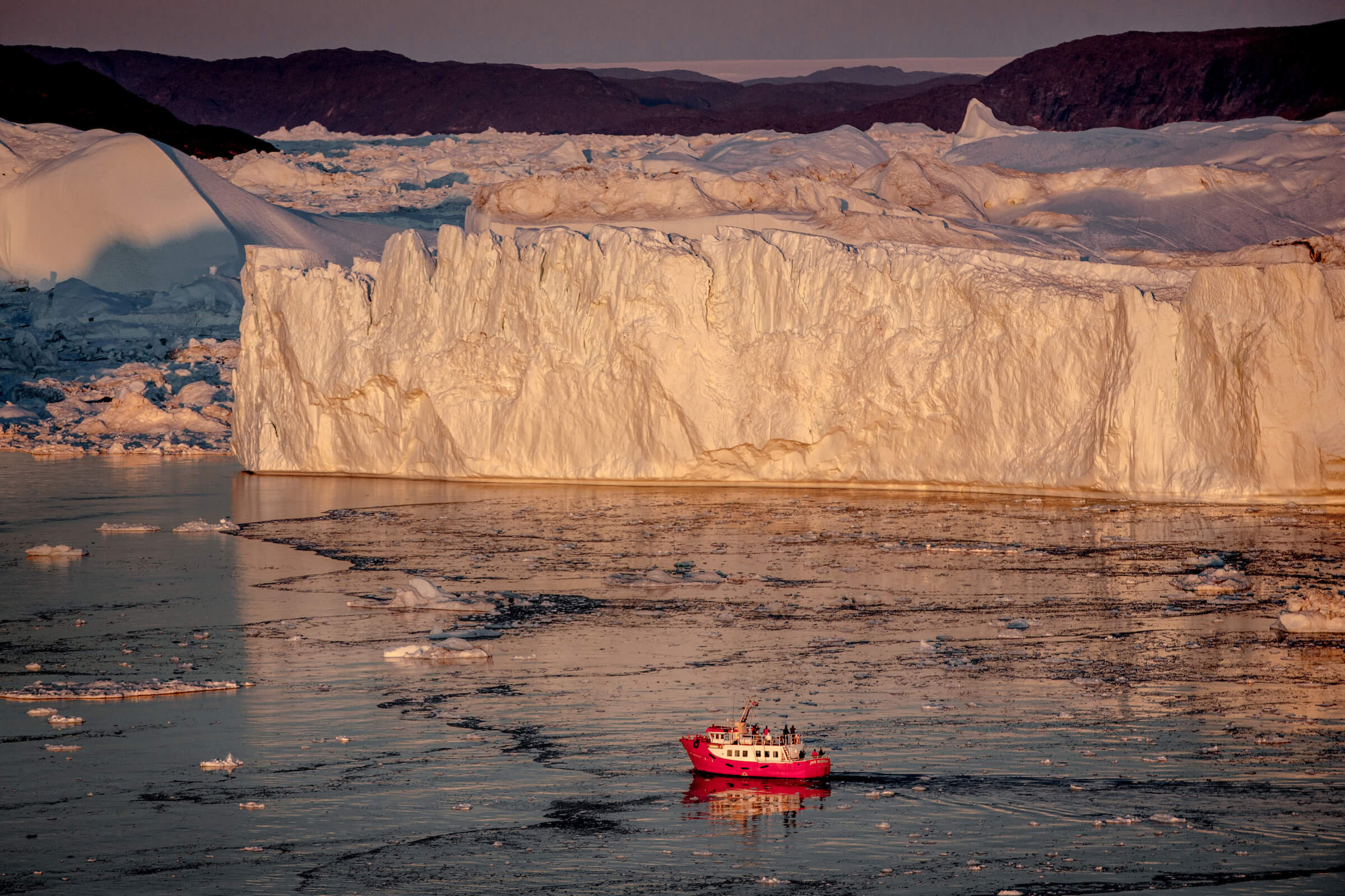 A passenger boat in the sunset near the icebergs of Ilulissat ice fjord in Greenland. By Mads Pihl