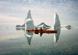 Two people kayaking next to large icebergs in crystal clear water. Photo by Arctic Dream